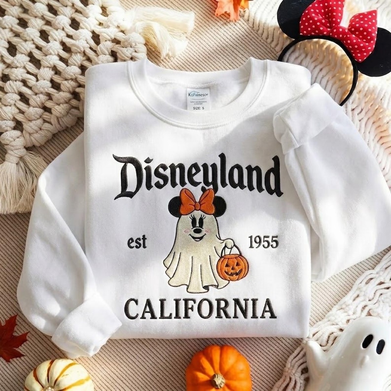 May include: A white crewneck sweatshirt featuring black embroidered text that reads Disneyland est 1955 CALIFORNIA. The center design shows a ghost character wearing black mouse ears with an orange bow, holding an orange jack-o'-lantern bucket.