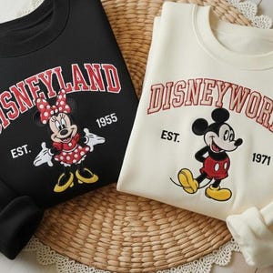 May include: Black sweatshirt with "Disneyland" in red arched text, featuring Minnie Mouse. A cream-colored sweatshirt displays "Disneyworld" with Mickey Mouse. Both sweatshirts have the establishment years 1955 and 1971.