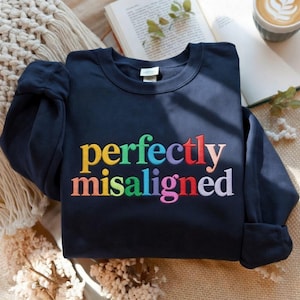 Op de afbeelding: Een marineblauwe sweatshirt met de geborduurde tekst "perfectly misaligned" in regenboogkleuren. De sweatshirt is opgevouwen, met de mouwen opgerold. Op de achtergrond zijn een boek, een kopje koffie en een crèmekleurige deken te zien.