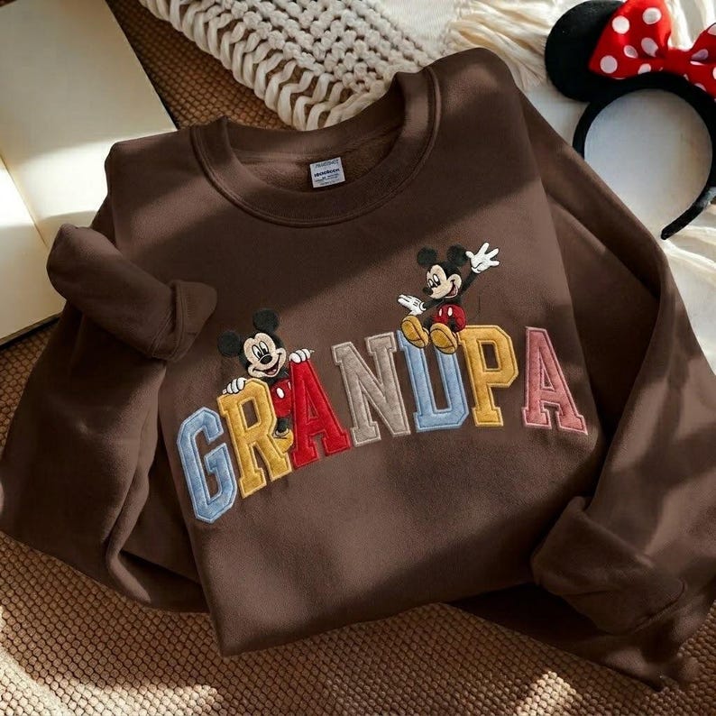 May include: A brown sweatshirt with the word "GRANDPA" in colourful, embroidered letters. Two cartoon mice are featured above the word. A Minnie Mouse ear headband is in the background.