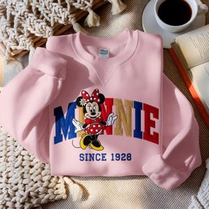 K&ouml;nnte beinhalten: Hellrosa Sweatshirt mit einem Minnie Mouse-Motiv. Das Motiv zeigt Minnie Mouse in einem roten gepunkteten Kleid und das Wort "MINNIE" in bunten Farben. Der Text "SINCE 1928" steht darunter. Das Sweatshirt ist gefaltet.