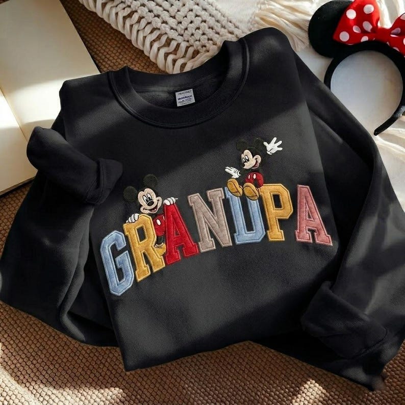 May include: Black sweatshirt featuring the word "GRANDPA" in colourful, embroidered letters. Mickey Mouse and a similar character are featured above the letters. A Minnie Mouse ear headband with a red bow is in the background.