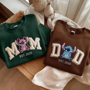Puede incluir: Dos sudaderas, una verde oscuro y otra marr&oacute;n. La verde tiene "MOM" con un personaje de dibujos animados rosa y "EST. 2025". La marr&oacute;n tiene "DAD" con un personaje azul y "EST. 2025".