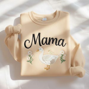 May include: A beige sweatshirt with the word "Mama" embroidered in black script. The design includes a white goose with a yellow beak and feet, a small yellow chick, and floral accents. The sleeves are folded.