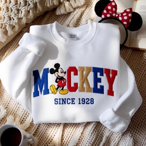 May include: White sweatshirt with the word "Mickey" in blue, red, and gold lettering, and a Mickey Mouse graphic. The text "SINCE 1928" is embroidered below. A red and white polka dot bow headband is visible.
