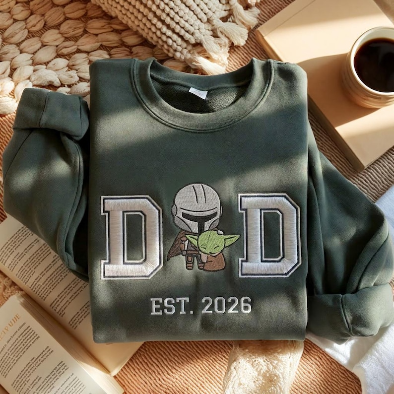 May include: Olive green sweatshirt with the word "DAD" in white embroidered letters, featuring an illustration of a Mandalorian and Grogu. The text "EST. 2026" is embroidered below. The sweatshirt is folded on a neutral surface.