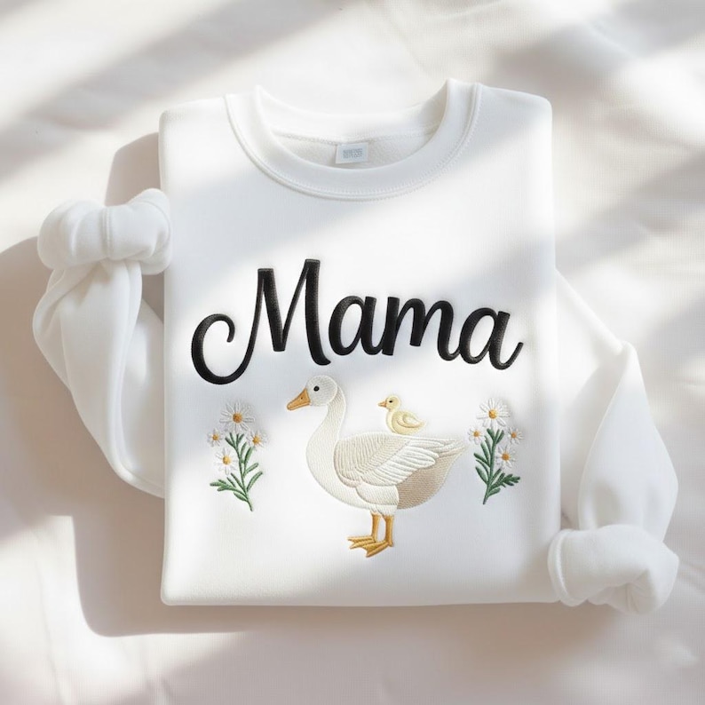 May include: White sweatshirt with the word "Mama" in black script. The sweatshirt features an embroidered design of a goose with a chick and floral accents. The sleeves are neatly folded, and the overall aesthetic is clean and simple.