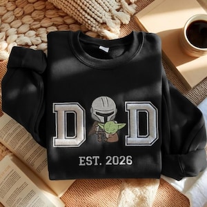 May include: Black sweatshirt with the word "DAD" in white and gray block letters, featuring an illustration of a Mandalorian and Grogu. The text "EST. 2026" is below the word "DAD".