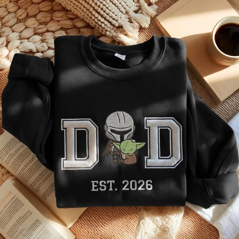 May include: Black sweatshirt with the word "DAD" in white and gray block letters, featuring an illustration of a Mandalorian and Grogu. The text "EST. 2026" is below the word "DAD".