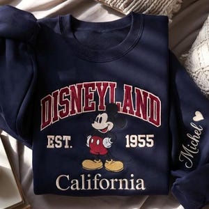 Puede incluir: Sudadera azul marino con la palabra "Disneyland" en un dise&ntilde;o arqueado granate, con "EST. 1955" debajo. Un gr&aacute;fico de Mickey Mouse est&aacute; centrado, y "California" est&aacute; impreso en la parte inferior. La manga tiene el nombre "Michel" y un coraz&oacute;n.