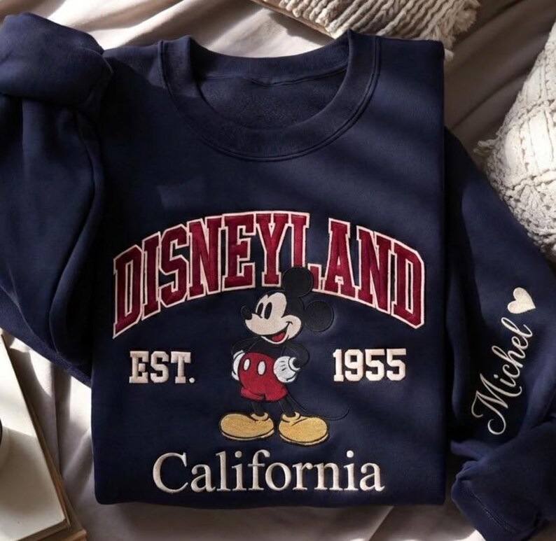 Puede incluir: Sudadera azul marino con la palabra "Disneyland" en un dise&ntilde;o arqueado granate, con "EST. 1955" debajo. Un gr&aacute;fico de Mickey Mouse est&aacute; centrado, y "California" est&aacute; impreso en la parte inferior. La manga tiene el nombre "Michel" y un coraz&oacute;n.