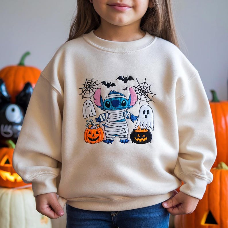 May include: Cream-colored sweatshirt featuring a Halloween-themed graphic. The design includes a blue alien wrapped in bandages, two ghosts, bats, and jack-o'-lanterns. The sweatshirt is a long-sleeved, crew neck style.