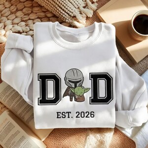 May include: White sweatshirt with the word "DAD" in black block letters, featuring a cartoon illustration of a Mandalorian character and Grogu. The text "EST. 2026" is below the illustration. The sweatshirt is folded on a light-colored surface.