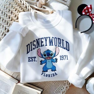 May include: White sweatshirt with the text "DISNEYWORLD EST. 1971 FLORIDA" in navy blue. The sweatshirt features an embroidered image of the character Stitch in blue. A black headband with Minnie Mouse ears and a red bow with white polka dots is also visible.