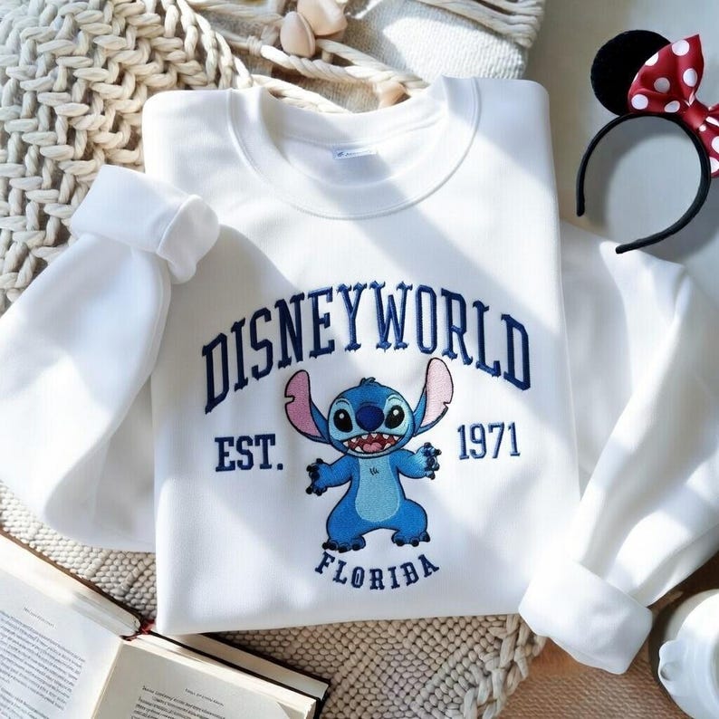 May include: White sweatshirt with the text "DISNEYWORLD EST. 1971 FLORIDA" in navy blue. The sweatshirt features an embroidered image of the character Stitch in blue. A black headband with Minnie Mouse ears and a red bow with white polka dots is also visible.