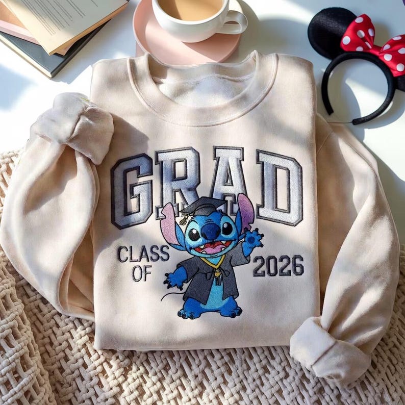 Puede incluir: Sudadera color crema con la palabra "GRAD" en letras grises, y "CLASS OF 2026" debajo. Un personaje de dibujos animados azul con toga de graduaci&oacute;n est&aacute; en el centro. Una diadema con orejas de Minnie Mouse est&aacute; en el fondo.
