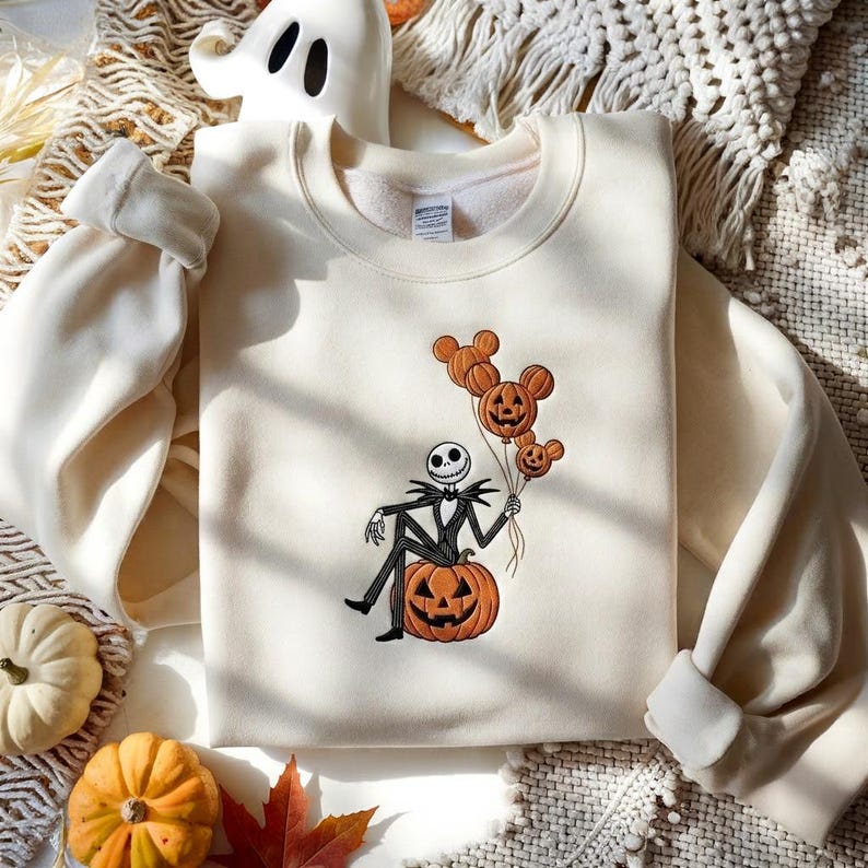 May include: A cream-colored crewneck sweatshirt featuring an embroidered graphic of a skeleton character sitting on a large orange jack-o'-lantern. The character holds three orange balloons shaped like mouse heads with jack-o'-lantern faces.