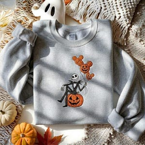 May include: A light gray crewneck sweatshirt featuring an embroidered graphic of a skeleton character sitting on a large orange pumpkin. The character holds a cluster of three orange pumpkin-shaped balloons with mouse-ear silhouettes.