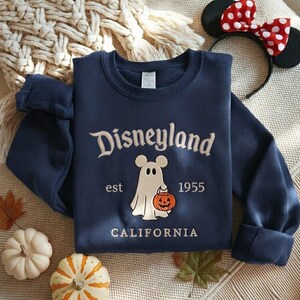 Pode incluir: Uma camisola azul-marinho de gola redonda com o texto bordado Disneyland est 1955 CALIFORNIA. O design central apresenta um fantasma branco com orelhas de rato a segurar uma ab&oacute;bora laranja com uma cara esculpida.