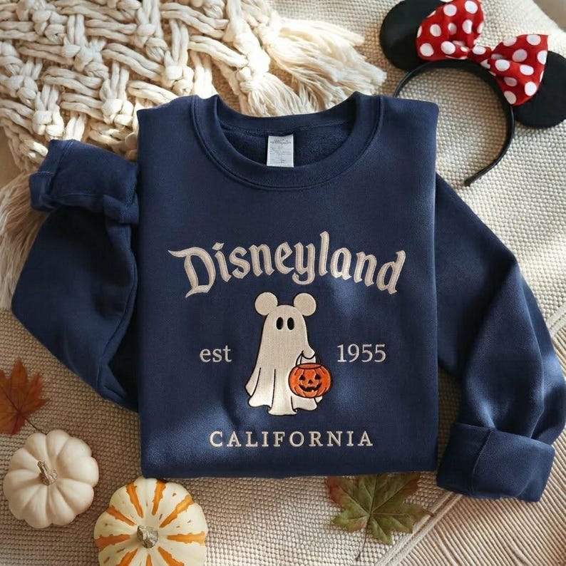 Pode incluir: Uma camisola azul-marinho de gola redonda com o texto bordado Disneyland est 1955 CALIFORNIA. O design central apresenta um fantasma branco com orelhas de rato a segurar uma ab&oacute;bora laranja com uma cara esculpida.