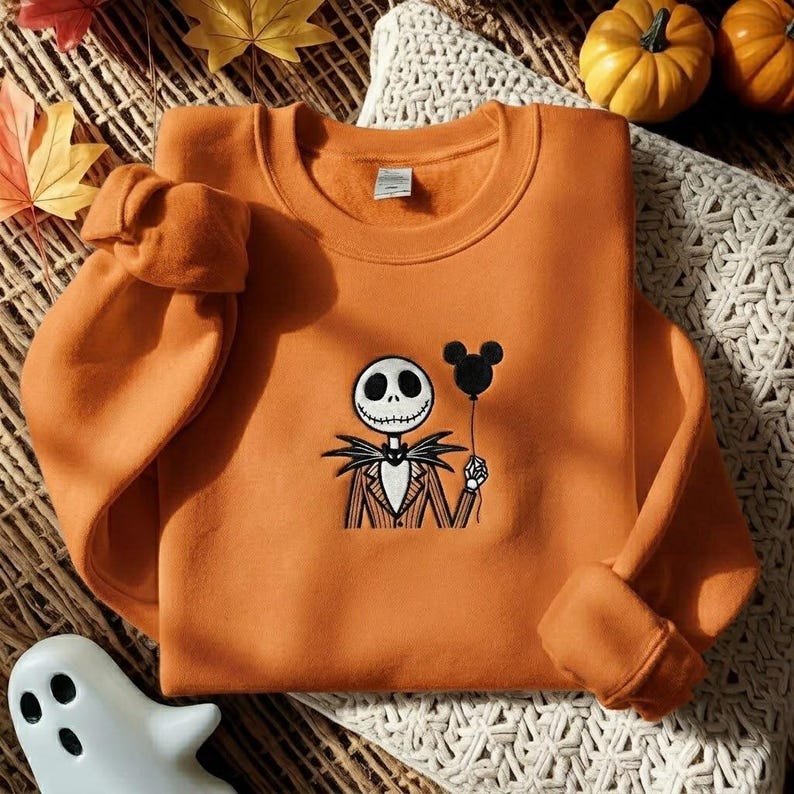 May include: An orange crewneck sweatshirt featuring an embroidered graphic of a skeletal character in a pinstriped suit holding a black balloon shaped like mouse ears. The design is centered on the chest of the long-sleeved pullover garment.