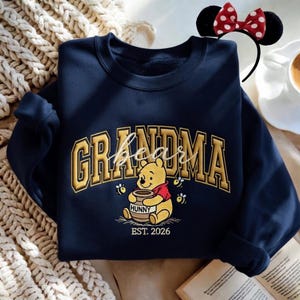 May include: Navy blue sweatshirt with the word "GRANDMA" in gold block letters, with "bear" in script. A Winnie the Pooh graphic holding a honey pot is below the text, with "HUNNY" and "EST. 2026". A Minnie Mouse ear headband is in the background.