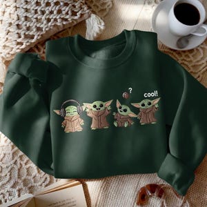May include: Dark green sweatshirt featuring a graphic of four cartoon characters. The characters are green with large ears and brown robes. One character wears headphones, and another has a question mark above its head. The word "cool!" is printed on the sweatshirt.