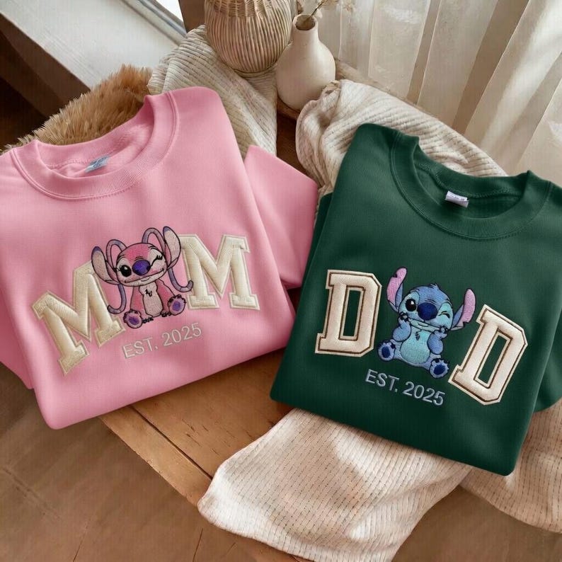Puede incluir: Dos sudaderas, una rosa y otra verde. La sudadera rosa tiene la palabra "MOM" bordada en letras color crema, con un personaje de dibujos animados y el texto "EST. 2025". La sudadera verde tiene "DAD" bordado con un personaje de dibujos animados y "EST. 2025".
