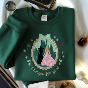 May include: A dark green sweatshirt with an embroidered design featuring two figures in front of a castle, surrounded by a scalloped border, bows, and stars. The text "Changed for good" is below the design.