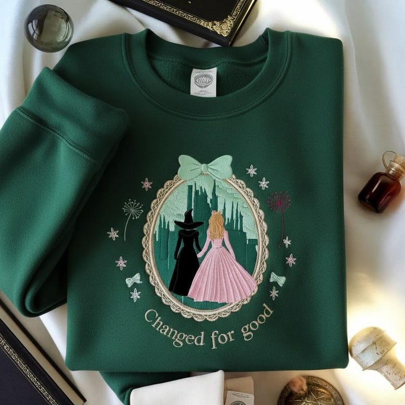 May include: A dark green sweatshirt with an embroidered design featuring two figures in front of a castle, surrounded by a scalloped border, bows, and stars. The text "Changed for good" is below the design.