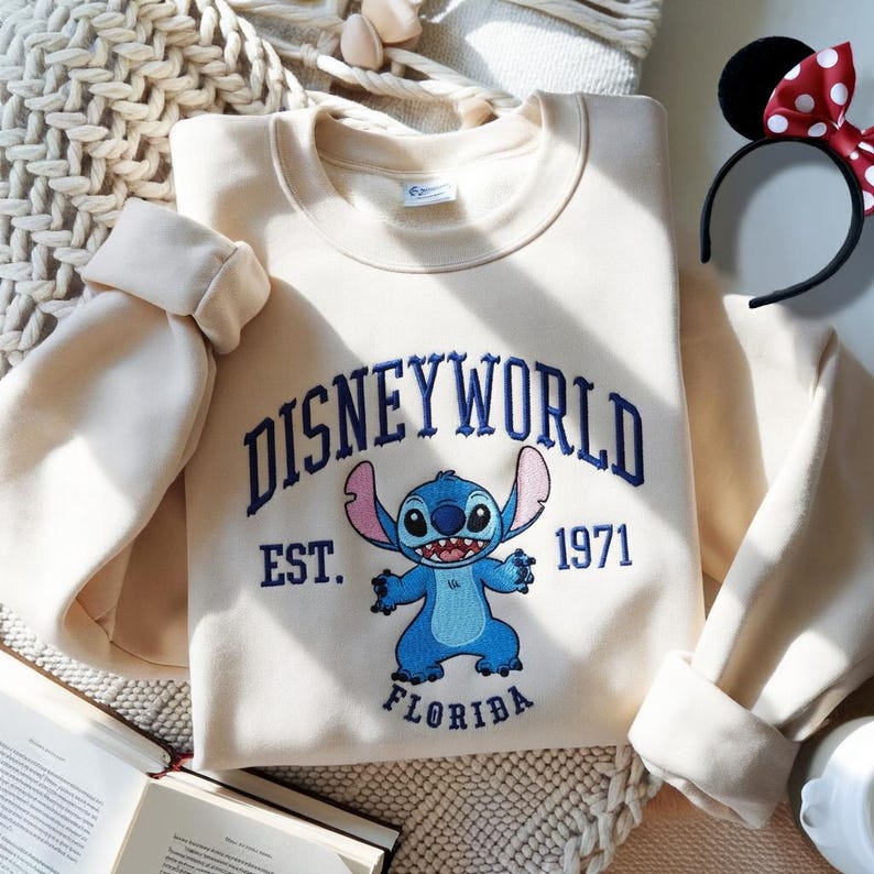 May include: Cream-colored sweatshirt with the text "DISNEYWORLD" embroidered in blue, along with a blue Stitch character and the text "EST. 1971 FLORIDA." A black headband with Minnie Mouse ears and a red and white polka dot bow is also visible.