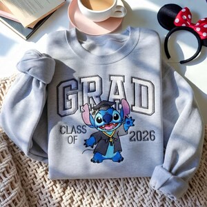 Puede incluir: Una sudadera gris claro con la palabra "GRAD" en una fuente universitaria, con "CLASS OF 2026" debajo. Un personaje de dibujos animados de Stitch con una toga de graduaci&oacute;n est&aacute; en el centro. Una diadema con orejas de Minnie Mouse est&aacute; en el fondo.