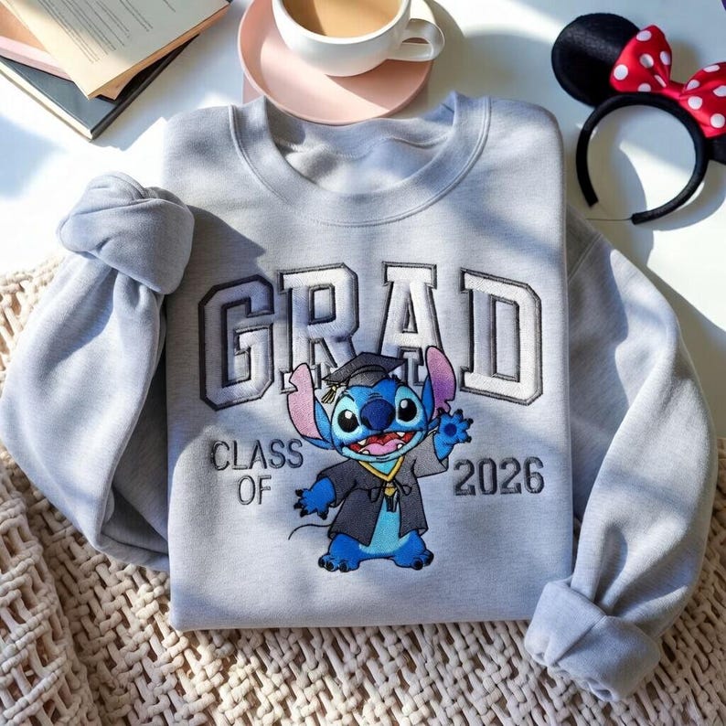 Puede incluir: Una sudadera gris claro con la palabra "GRAD" en una fuente universitaria, con "CLASS OF 2026" debajo. Un personaje de dibujos animados de Stitch con una toga de graduaci&oacute;n est&aacute; en el centro. Una diadema con orejas de Minnie Mouse est&aacute; en el fondo.