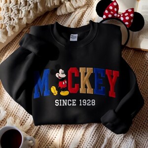 May include: Black sweatshirt with the word "Mickey" in blue, red, and gold embroidery, featuring Mickey Mouse and the text "Since 1928". A Minnie Mouse ear headband with a red and white polka dot bow is visible.