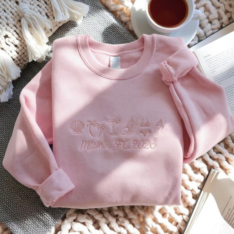 Peut inclure: Un sweat-shirt rose clair avec des ic&ocirc;nes brod&eacute;es sur le th&egrave;me de la plage et le texte "Miami, FL 2026". Le motif comprend un coquillage, un palmier, un cocktail, un bikini et une glace. Le sweat-shirt est pli&eacute; sur une surface textur&eacute;e.