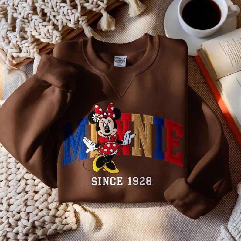 K&ouml;nnte beinhalten: Ein braunes Sweatshirt mit Minnie Mouse und dem Wort "MINNIE" in Blau, Gold und Rot. Minnie Mouse tr&auml;gt ein rotes gepunktetes Kleid und eine Schleife. Der Text "SINCE 1928" steht darunter. Das Sweatshirt liegt auf einer strukturierten Oberfl&auml;che.