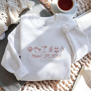 Peut inclure: Sweat-shirt blanc avec broderie corail de motifs estivaux : coquillage, palmier, cocktail et bikini. Le texte "Miami, FL, 2026" est brod&eacute; en dessous. Le sweat-shirt est pli&eacute; sur une surface textur&eacute;e.