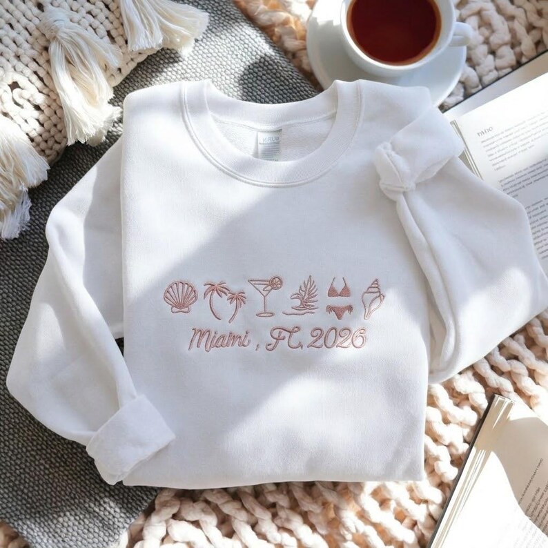 Peut inclure: Sweat-shirt blanc avec broderie corail de motifs estivaux : coquillage, palmier, cocktail et bikini. Le texte "Miami, FL, 2026" est brod&eacute; en dessous. Le sweat-shirt est pli&eacute; sur une surface textur&eacute;e.