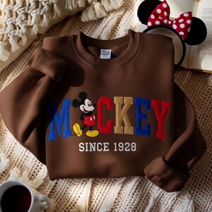 May include: A brown sweatshirt with the word "Mickey" embroidered in blue, red, and gold, and a cartoon Mickey Mouse graphic. The text "SINCE 1928" is below. A Mickey Mouse ear headband with a red and white polka dot bow is also visible.