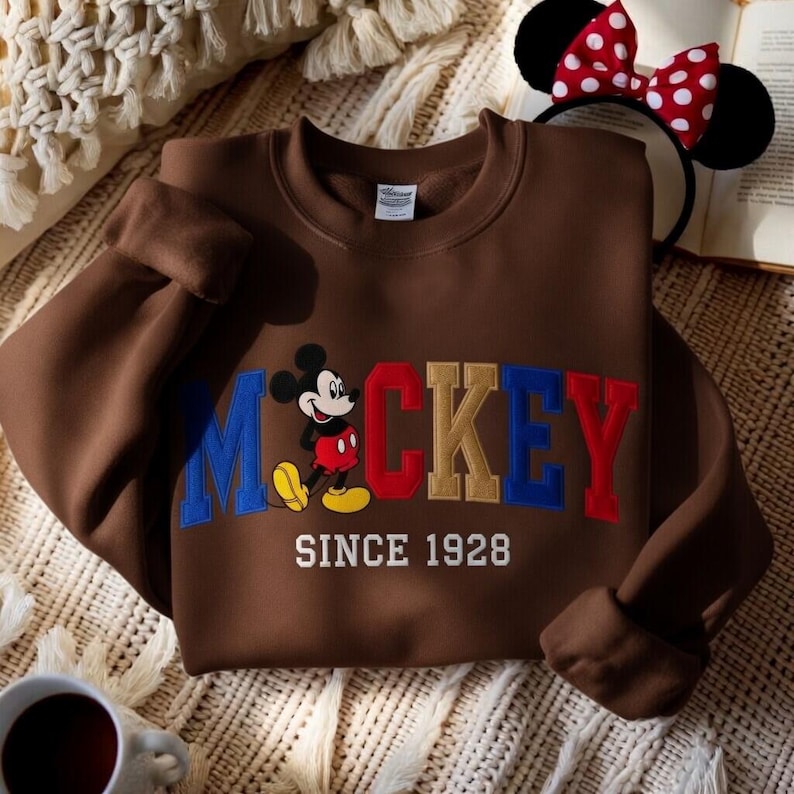May include: A brown sweatshirt with the word "Mickey" embroidered in blue, red, and gold, and a cartoon Mickey Mouse graphic. The text "SINCE 1928" is below. A Mickey Mouse ear headband with a red and white polka dot bow is also visible.