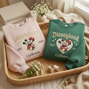 May include: Two sweatshirts, one pink and one green, are displayed in a woven basket. Both feature the embroidered word "Disneyland" in gold script, along with a heart-shaped graphic of Mickey and Minnie Mouse. The text "EST 1955 California" is also embroidered.