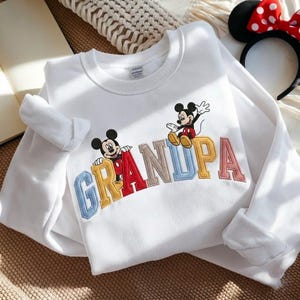 May include: White sweatshirt with the word "GRANDPA" embroidered in colourful letters, featuring Mickey Mouse cartoon characters. The sweatshirt has long sleeves and a crew neck. A Minnie Mouse ear headband with a red bow is in the background.