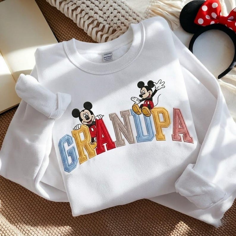 May include: White sweatshirt with the word "GRANDPA" embroidered in colourful letters, featuring Mickey Mouse cartoon characters. The sweatshirt has long sleeves and a crew neck. A Minnie Mouse ear headband with a red bow is in the background.