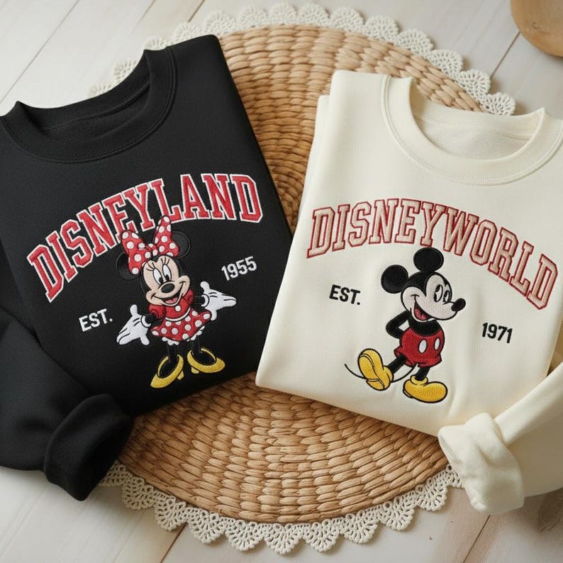May include: Two sweatshirts, one black and one cream-colored, featuring Disney characters. The black sweatshirt displays "Disneyland" with Minnie Mouse, and the cream one says "Disneyworld" with Mickey Mouse. Both have the establishment year.