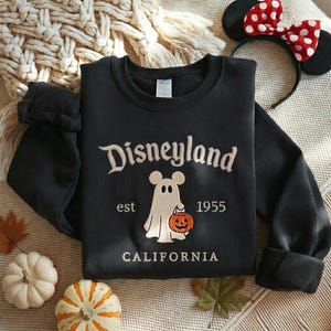 Pode incluir: Uma camisola preta de manga comprida com o texto bordado Disneyland est 1955 CALIFORNIA. O design central apresenta um fantasma branco com orelhas de rato a segurar uma ab&oacute;bora cor de laranja com uma cara esculpida.