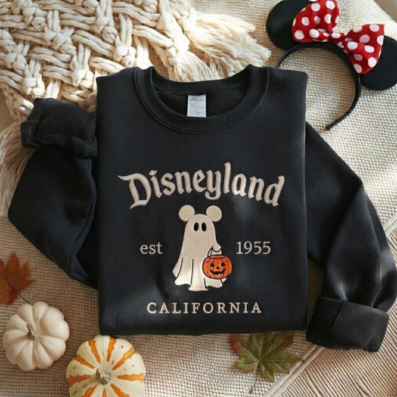 Pode incluir: Uma camisola preta de manga comprida com o texto bordado Disneyland est 1955 CALIFORNIA. O design central apresenta um fantasma branco com orelhas de rato a segurar uma ab&oacute;bora cor de laranja com uma cara esculpida.