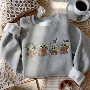 May include: A gray sweatshirt with white cuffs featuring a cartoon design of four Grogu characters. One Grogu wears headphones, another has arms raised, a third is seated, and the last says "cool!" above it.