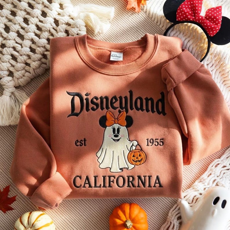 May include: A terracotta-colored crewneck sweatshirt featuring black embroidered text that reads Disneyland California est 1955. The center graphic depicts a white ghost character wearing a black and orange bow, holding a small orange jack-o'-lantern.