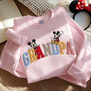 May include: A pale pink sweatshirt with the word "GRANDPA" embroidered in various colours. Two Mickey Mouse cartoon characters are positioned above the letters. A black headband with a red bow is in the background.