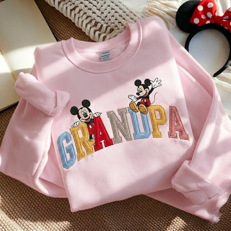 May include: A pale pink sweatshirt with the word "GRANDPA" embroidered in various colours. Two Mickey Mouse cartoon characters are positioned above the letters. A black headband with a red bow is in the background.